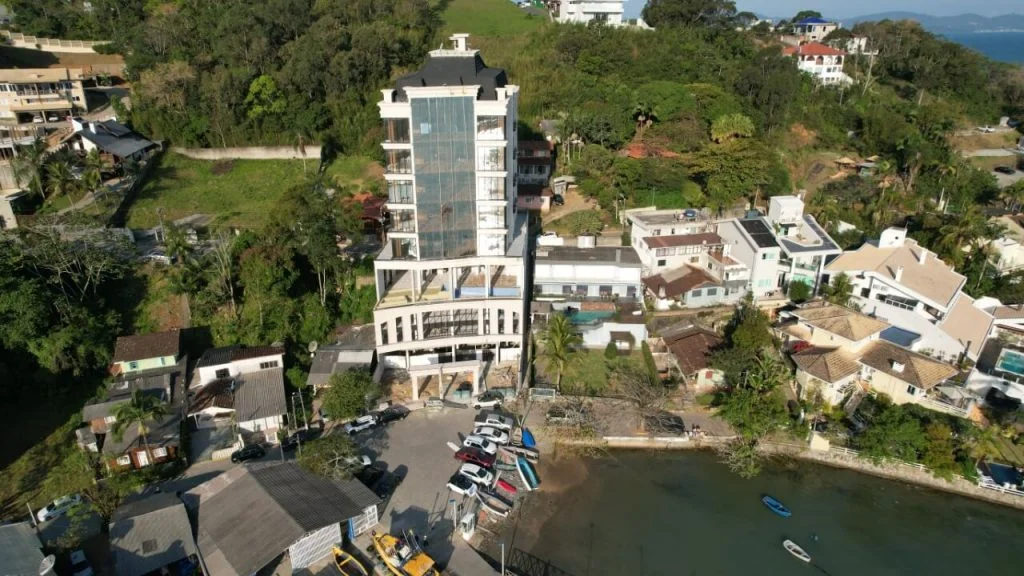 Imagem de um edifício moderno de vários andares próximo a uma área de água, com várias casas ao redor e muita vegetação ao fundo, em uma área cercada por natureza em Itapema