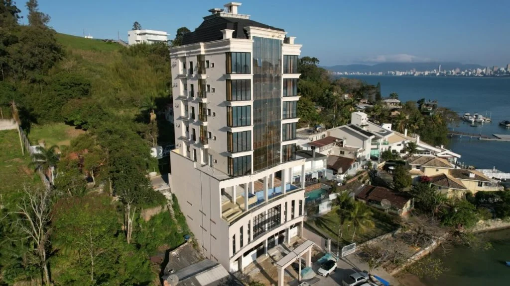 Imagem de um edifício residencial moderno localizado na orla de uma cidade à beira-mar, com vista para o mar e bairro residencial ao redor, na cidade de Itapema
